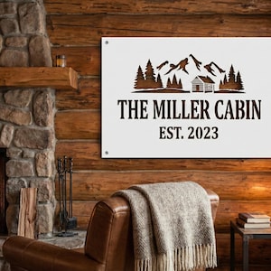May include: A white metal sign with the text "THE MILLER CABIN EST. 2023" and a mountain and cabin design in brown. The sign is mounted on a wooden wall above a leather armchair with a blanket.