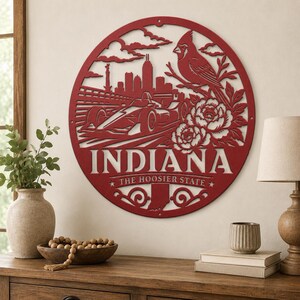 Może przedstawiać: Czerwony metalowy obraz na ścianę z napisem "INDIANA" i frazą "THE HOOSIER STATE". Projekt zawiera samoch&oacute;d wyścigowy, panoramę miasta, ptaka kardynała i kwiaty, wszystko w okrągłej ramie.