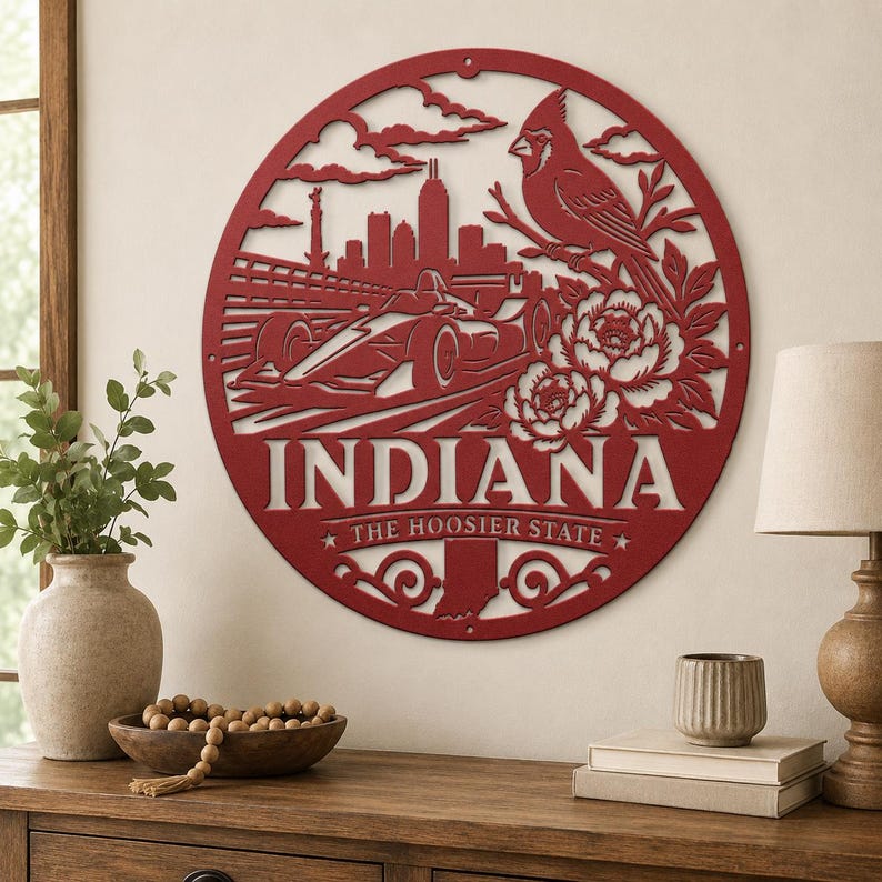 Może przedstawiać: Czerwony metalowy obraz na ścianę z napisem "INDIANA" i frazą "THE HOOSIER STATE". Projekt zawiera samoch&oacute;d wyścigowy, panoramę miasta, ptaka kardynała i kwiaty, wszystko w okrągłej ramie.