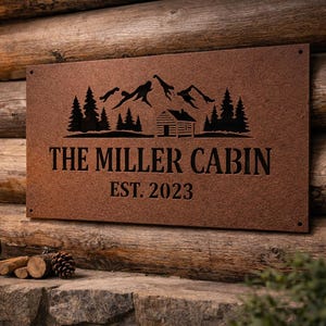 May include: A rectangular, brown metal sign with the text "THE MILLER CABIN" and "EST. 2023" in black. The sign features a mountain and cabin silhouette design. The sign is mounted on a wooden cabin wall.