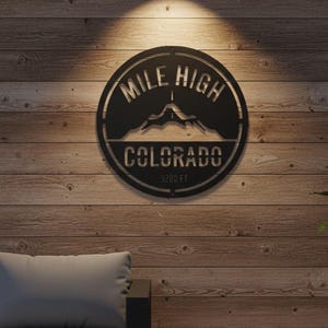 May include: A black metal wall art piece with the words "Mile High Colorado" and a mountain silhouette. The text "5280 FT" is below the word "Colorado". The art is circular and mounted on a wooden wall.