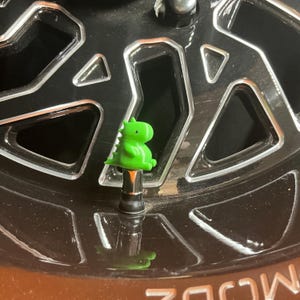 May include: A green dinosaur-shaped valve stem cap is mounted on a black and silver rim. The dinosaur has a friendly, cartoonish design. The rim has a geometric pattern with triangular and rectangular shapes.