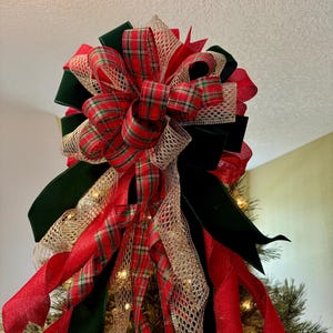 May include: A festive Christmas tree topper featuring a large bow made of red plaid, green velvet, and gold mesh ribbons. The bow is layered and has long, flowing red ribbons. The tree is decorated with small white lights.