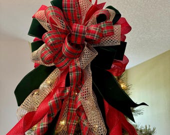 May include: A festive Christmas tree topper featuring a large bow made of red plaid, green velvet, and gold mesh ribbons. The bow is layered and has long, flowing red ribbons. The tree is decorated with small white lights.