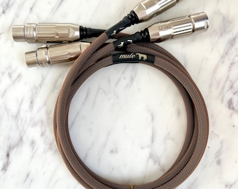 May include: Two brown braided audio cables with silver XLR connectors. The cables are wrapped in a brown mesh and have a black label with the word "mule" and a small elephant graphic.