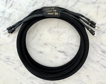 May include: Four black speaker cables with gold lettering that reads "mule" on each cable. The cables are coiled together on a white marble surface.