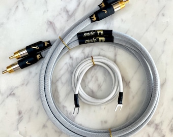 May include: Audio cables with gold-colored connectors and black bands with the word "mule" printed on them. The cables are gray and white, with a woven texture. One set is coiled, and another set is straight.