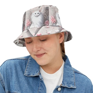 May include: A white bucket hat with a pink and white cat print. The hat has a wide brim and a flat crown. The cat print features a white cat with pink flowers and a white background.
