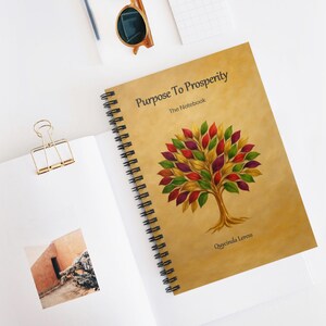 Puede incluir: Un cuaderno con espiral con el título "Purpose To Prosperity" y una ilustración de un árbol con hojas coloridas. El cuaderno tiene una cubierta de color beige y encuadernación en espiral negra. El cuaderno está abierto sobre una superficie blanca.