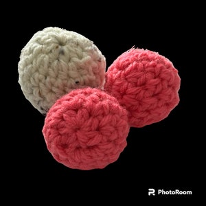 Puede incluir: Tres bolas de ganchillo hechas a mano en diferentes tonos de rosa y blanco roto. Las bolas son redondas y texturizadas, con patrones de puntadas visibles. La bola blanca rota tiene motas de colores más oscuros. Probablemente son decorativas o para jugar.