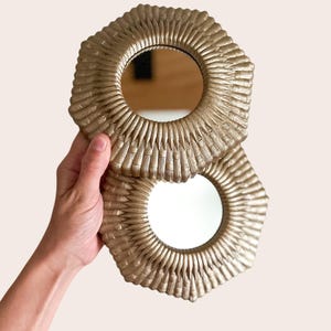 May include: Two circular mirrors with textured gold frames featuring a radiating sunburst pattern of vertical ridges. The frames have a metallic finish and surround a central reflective glass surface. The mirrors are stacked slightly overlapping.