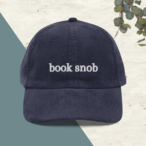 May include: A navy blue corduroy baseball cap with white embroidery that reads "book snob".