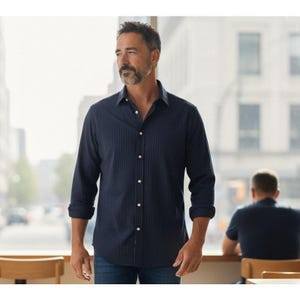 May include: A navy blue button-down shirt with vertical stripes. The long-sleeved shirt has white buttons and is paired with blue jeans. The sleeves are rolled up. The person is standing near a window.