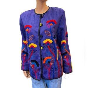 May include: A purple jacket with a floral design. The jacket has a textured pattern and is adorned with colorful embroidered flowers in red, yellow, blue, and orange. It features black buttons and trim.