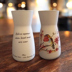 May include: Two white ceramic vases with a textured finish. One vase has a robin and flowers printed on it, the other has the text "Robins appear when loved ones are near.  Mom 21st June 1925 - 27th December 2021"