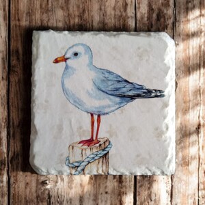May include: A white and blue watercolor painting of a seagull perched on a wooden post with a blue rope. The seagull has orange legs and a yellow beak.