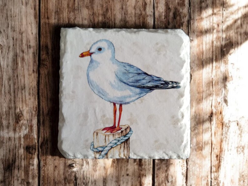 May include: A white and blue watercolor painting of a seagull perched on a wooden post with a blue rope. The seagull has orange legs and a yellow beak.