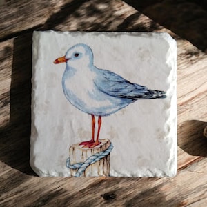 May include: A white ceramic coaster with a watercolor painting of a blue and white seagull perched on a wooden post with a blue rope. The seagull has orange legs and a yellow beak.