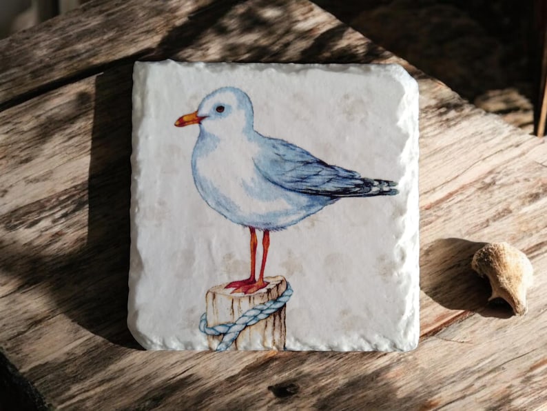 May include: A white ceramic coaster with a watercolor painting of a blue and white seagull perched on a wooden post with a blue rope. The seagull has orange legs and a yellow beak.