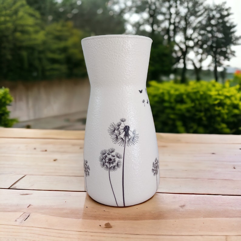 May include: White ceramic vase with a textured surface and a black and white dandelion design. The vase has a narrow neck and a wide base.