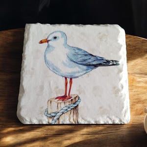 May include: A white ceramic coaster with a watercolor painting of a blue and white seagull perched on a wooden post with a blue rope. The seagull has red legs and a yellow beak.