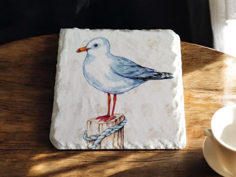 May include: A white ceramic coaster with a watercolor painting of a blue and white seagull perched on a wooden post with a blue rope. The seagull has red legs and a yellow beak.