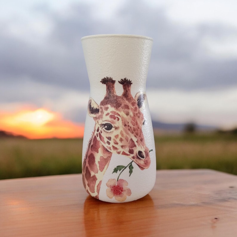 May include: White ceramic vase with a hand-painted giraffe design. The giraffe is brown and has a pink flower in its mouth. The vase is perfect for displaying fresh flowers.