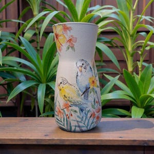 May include: A cream ceramic vase with a flared top, decorated with a painted design of two parakeets on a branch with flowers. The vase has a textured surface and is set against a backdrop of green plants.