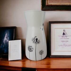 May include: A white ceramic vase with a textured surface and a design of black dandelions. The vase is sitting on a wooden surface with two picture frames.