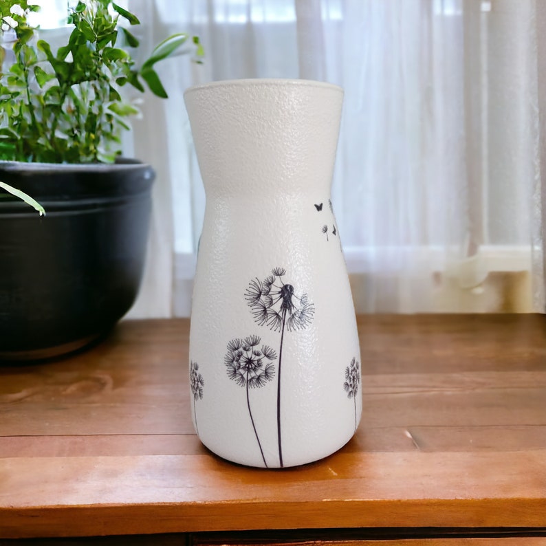 May include: White ceramic vase with a textured surface and a black and white dandelion design. The vase has a narrow neck and a wide base.