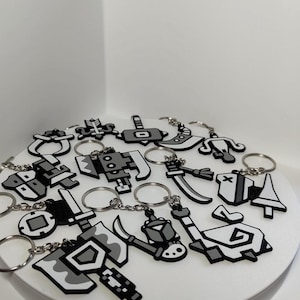 Könnte beinhalten: Eine Sammlung von schwarzen, weißen und grauen Schlüsselanhängern mit verschiedenen Waffen- und Objekt-Designs. Jeder Schlüsselanhänger hat einen silberfarbenen Ring. Die Schlüsselanhänger sind auf einer weißen Oberfläche angeordnet.