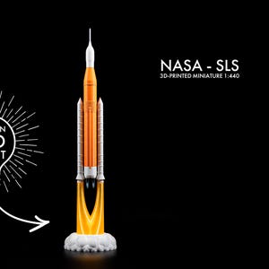 Peut inclure: Maquette miniature imprimée en 3D d'une fusée NASA SLS avec lumière LED intégrée. La fusée est orange et blanche, avec un effet de flamme à la base. Le texte "NASA - SLS 3D-PRINTED MINIATURE 1:440" est affiché.