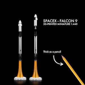 Peut inclure: Miniature imprimée en 3D de la fusée SpaceX Falcon 9, échelle 1:440, avec un corps blanc et noir et une base orange. Le texte indique "SPACEX - FALCON 9 3D-PRINTED MINIATURE 1:440". La fusée est aussi épaisse qu'un crayon.