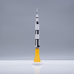 May include: A detailed model of a Saturn V rocket, painted in white, black, and silver, with yellow flames and white smoke emanating from the base. The rocket is standing upright, showcasing its multi-stage design, perfect for space enthusiasts.