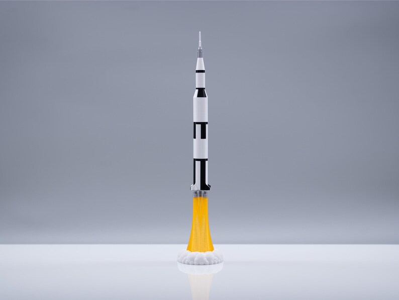 May include: A detailed model of a Saturn V rocket, painted in white, black, and silver, with yellow flames and white smoke emanating from the base. The rocket is standing upright, showcasing its multi-stage design, perfect for space enthusiasts.