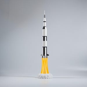 May include: A model rocket in a vertical position, featuring a white and black body with a pointed top. The rocket is depicted with yellow flames emanating from its base, resting on a white cloud-like structure. The overall design suggests a space exploration theme.