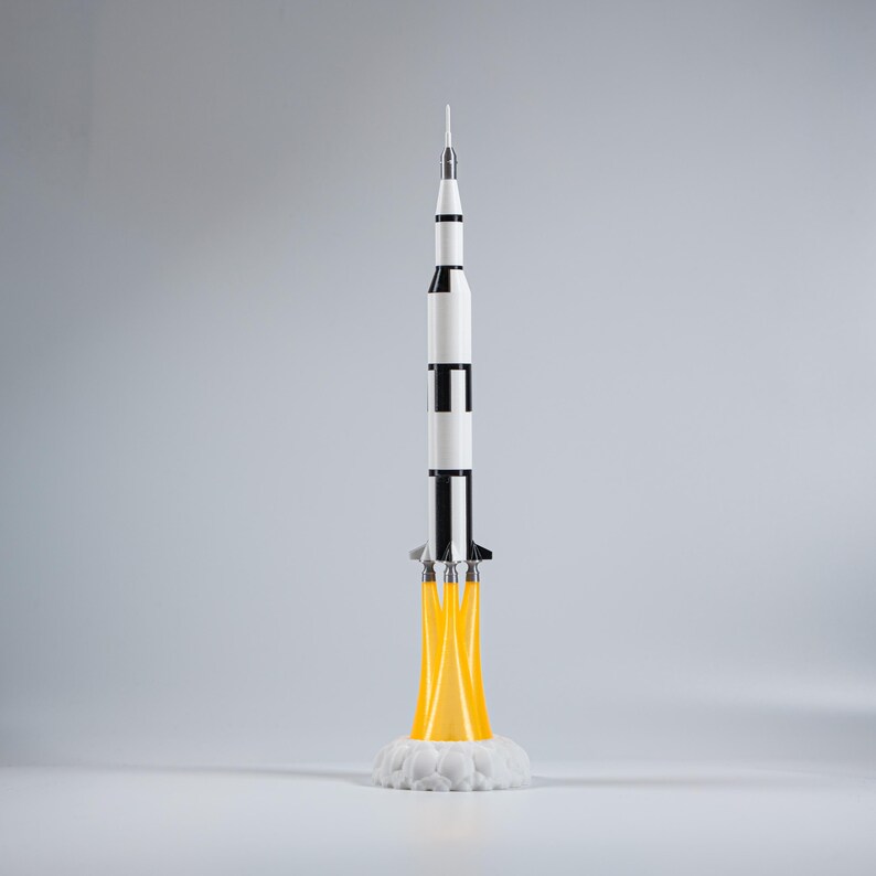 May include: A model rocket in a vertical position, featuring a white and black body with a pointed top. The rocket is depicted with yellow flames emanating from its base, resting on a white cloud-like structure. The overall design suggests a space exploration theme.