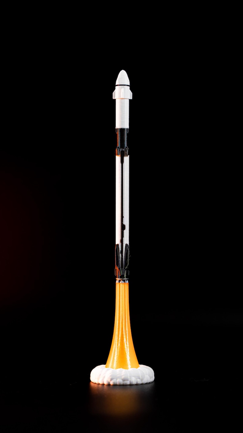 May include: A vertical image of a toy rocket ship against a black background. The rocket has a white nose cone, a white and black body, and an orange flame effect at the base, sitting on a white base.