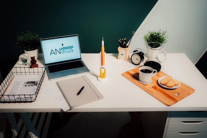 May include: A desk setup featuring a laptop with "ANdruck" on the screen, a rocket model, a notebook, and a coffee cup with biscuits on a wooden board. A black wire basket holds papers, and a small potted plant is visible.