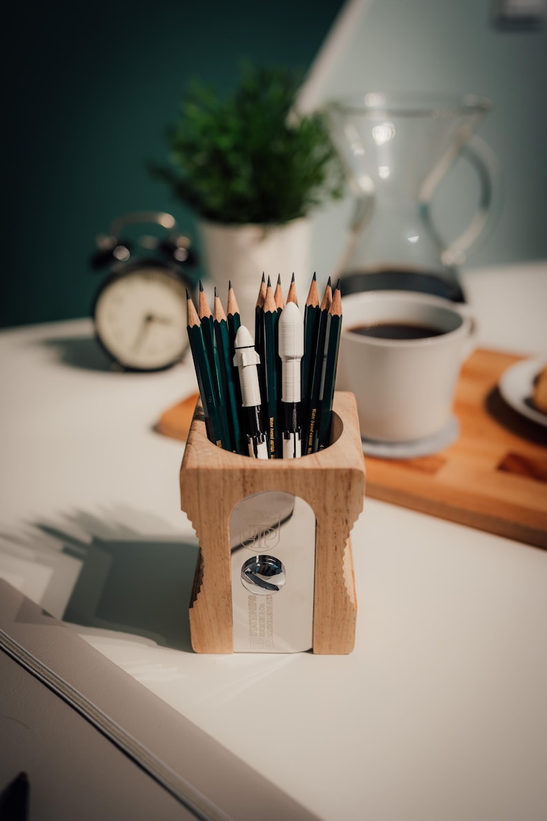 May include: Wooden pencil sharpener with pencils inside. The sharpener has a silver blade and a rectangular shape. The pencils are green and black. A white mug of coffee and a small plant are in the background.