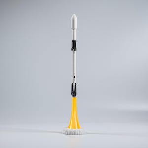 Op de afbeelding: Een speelgoedraket met een witte neuskegel, een zilveren romp en zwarte accenten. De raket heeft een gele vlammenbasis met witte rook. De raket staat rechtop tegen een grijze achtergrond.