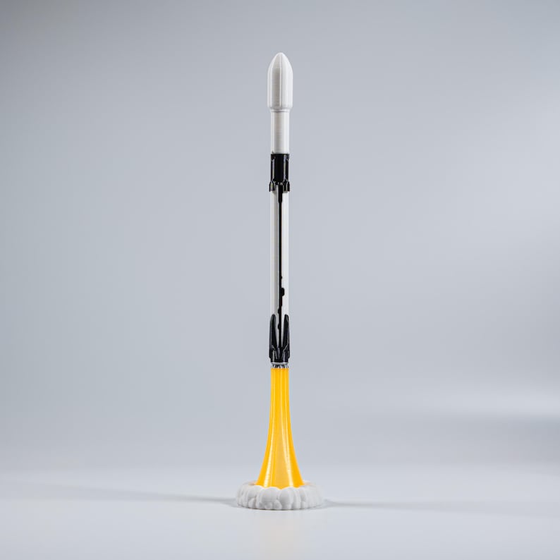 Op de afbeelding: Een speelgoedraket met een witte neuskegel, een zilveren romp en zwarte accenten. De raket heeft een gele vlammenbasis met witte rook. De raket staat rechtop tegen een grijze achtergrond.