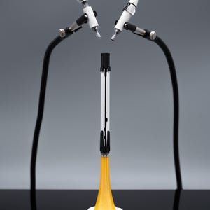May include: A white and black rocket model with a yellow flame base, set on a white cloud-like base. Two white and silver objects are held by black flexible arms. The background is a gradient of gray.
