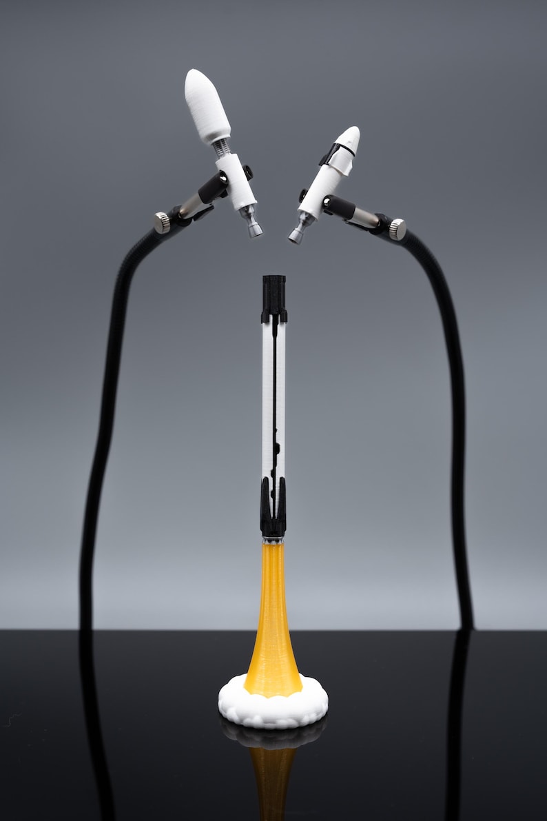May include: A white and black rocket model with a yellow flame base, set on a white cloud-like base. Two white and silver objects are held by black flexible arms. The background is a gradient of gray.