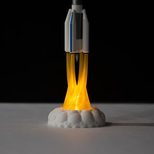 May include: A miniature rocket ship model with a white and blue body, emitting a bright yellow glow from its base, simulating flames. The rocket sits on a white, cloud-like base, creating a dynamic and illuminated display. The background is black.