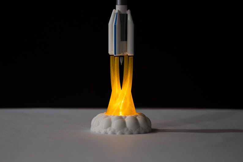 May include: A miniature rocket ship model with a white and blue body, emitting a bright yellow glow from its base, simulating flames. The rocket sits on a white, cloud-like base, creating a dynamic and illuminated display. The background is black.