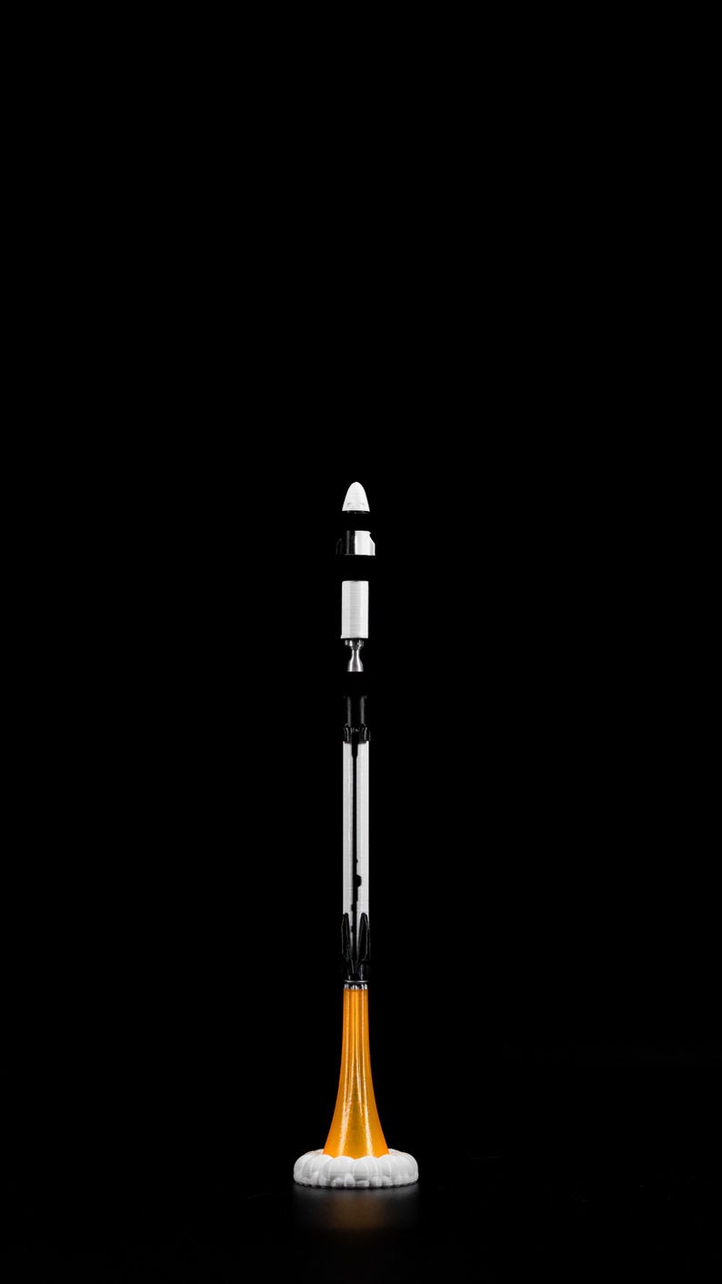 May include: A white and black model rocket with an orange and white base, set against a black background. The rocket is segmented, with a conical top and a detailed engine section. The base emits a stylized orange flame.