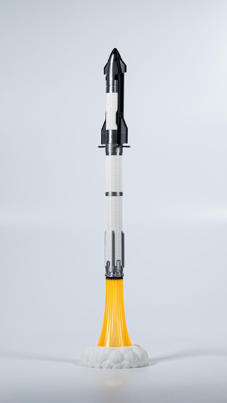May include: A model rocket with a black and silver nose cone, a white cylindrical body, and silver accents. The rocket sits on a white cloud-shaped base with orange flames emanating from the bottom, suggesting liftoff.