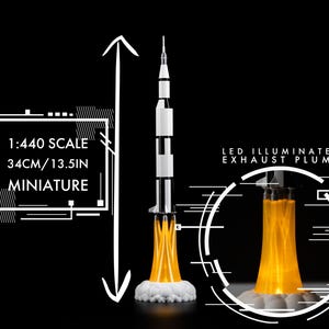 May include: A miniature model rocket, 13.5 inches tall, with a black and white body and an LED-illuminated exhaust plume. The model is 1:440 scale and sits on a white cloud-like base. The background is black.