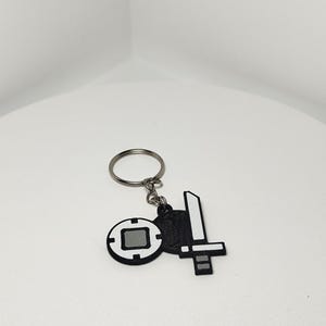 May include: A black and white keychain featuring two charms: a sword and a circular emblem. The sword charm has a white blade and a black handle. The circular emblem has a gray square in the center and a black border.